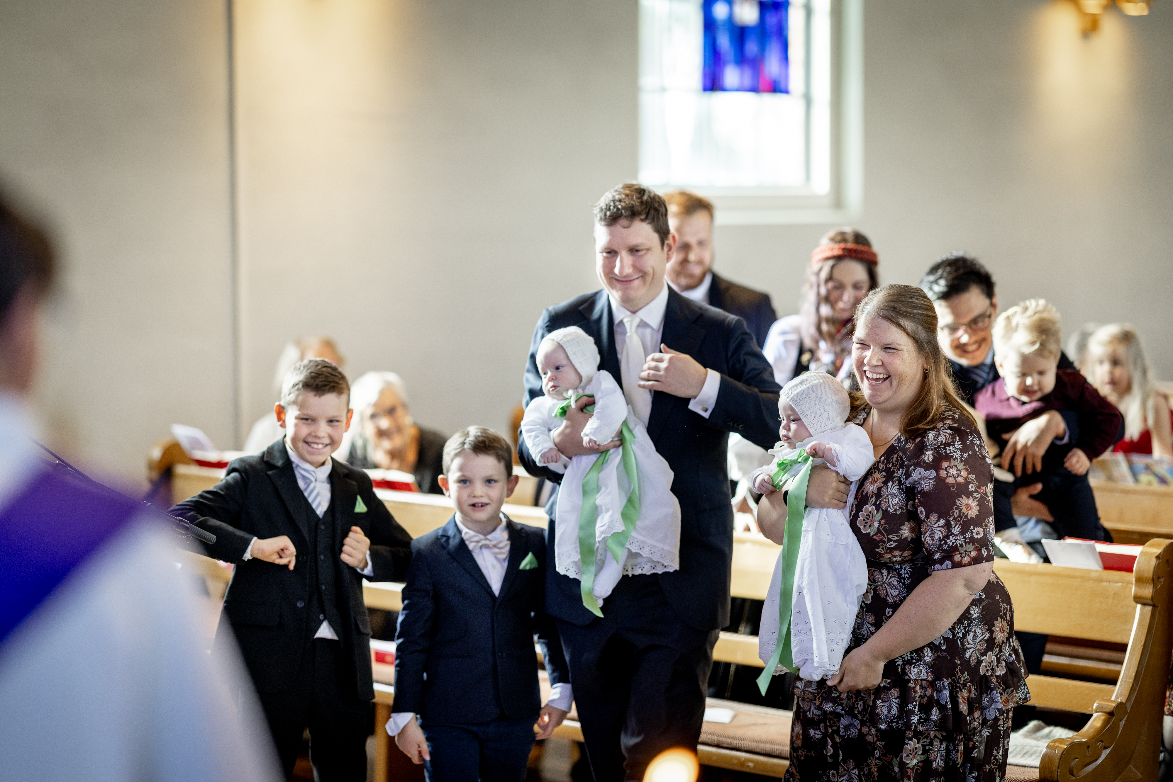 Mor og far med tvillinggutter i dåpskjoler, og storebrødrene ved siden av seg reiser seg fra kirkebenken klare til å gå opp til døpefonten.